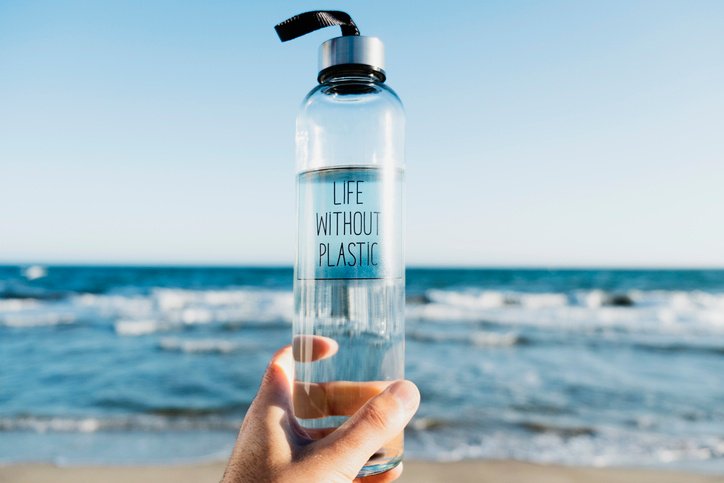 Life without plastic written on a clear reusable water bottle. The water bottle is held at the beach overlooking the ocean