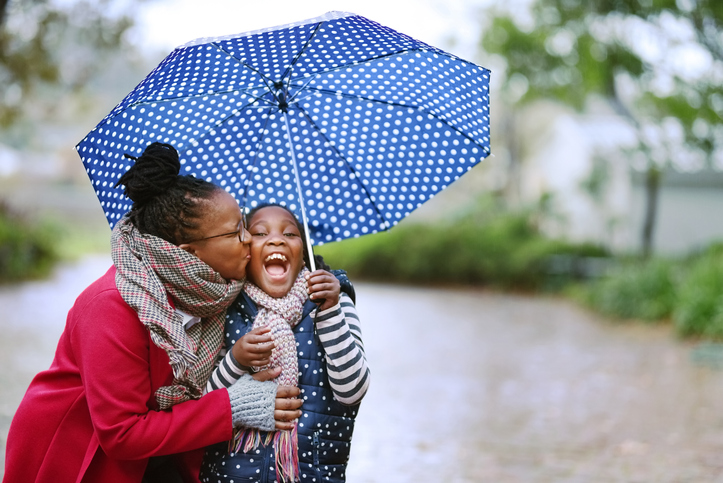 Picture of a black woman kissing her daughter while hiding from the rain under an umbrella