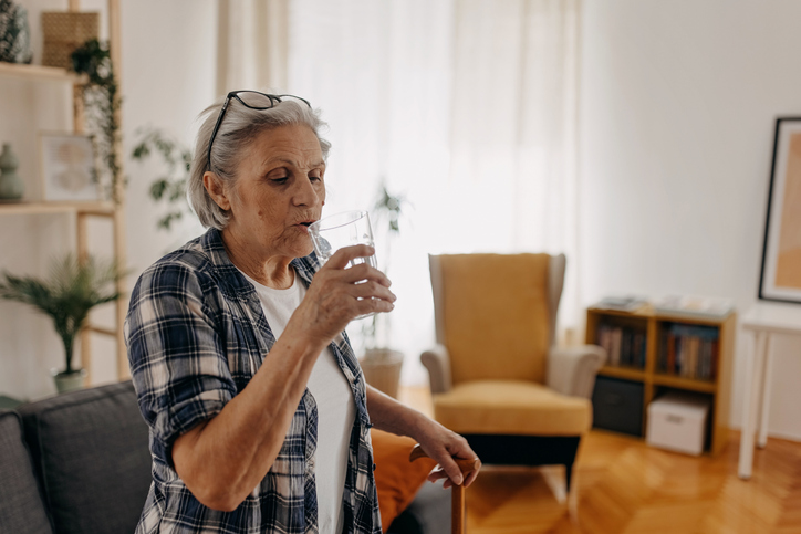 Senior woman is thirsty, drinking glass of water, holding on walking stick