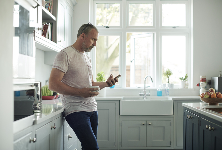 mature man standing in kitchen, scrolling on phone,