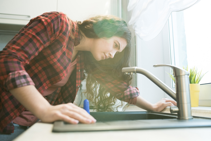 Serious concentrated young woman in checkered shirt checking faucet while having problem with dropping tap in kitchen