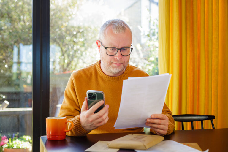 Adult man dressed wearing a yellow sweater and jeans while in kitchen at home checking his bills