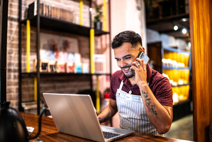 Small business owner young man using laptop and talking on mobile phone at a coffee shop