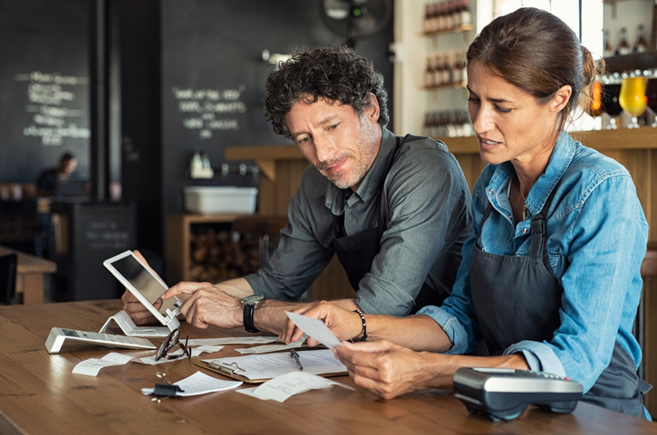 Man and woman sitting in cafeteria discussing finance for the month. Stressed couple looking at bills sitting in restaurant wearing uniform apron. Café staff sitting together looking at expenses and bills