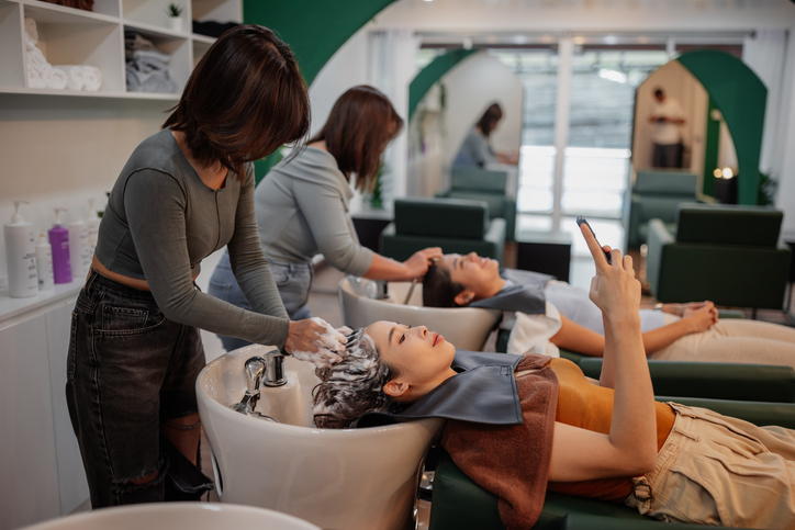 Smiling woman is using a phone while hairdresser is washing her hair at salon.