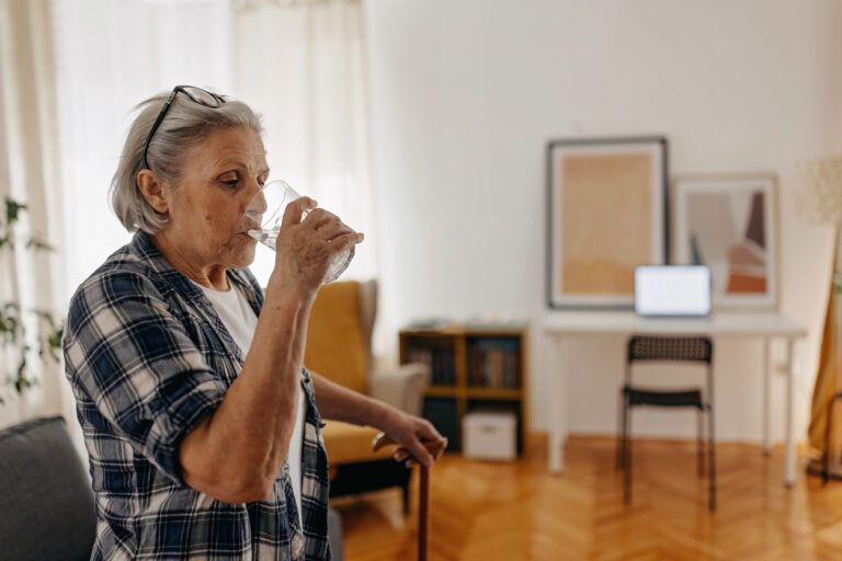 An elderly lady is drinking a glass of water in her home, while holding onto a walking stick with the other hand.