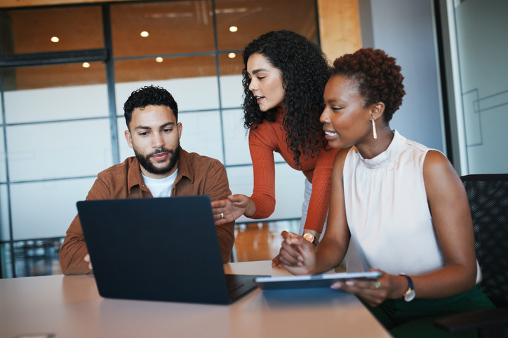 Three diverse professionals collaborate around a laptop in a modern office. Two women and a man discuss content on the screen, with one woman pointing.